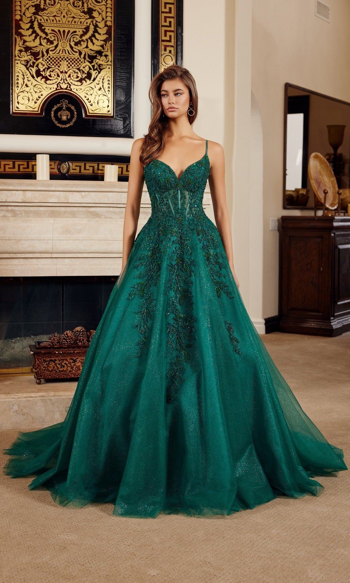 A woman stands indoors wearing the Nox Anabel H1883, a floor-length emerald green prom dress with a fitted corset bodice, intricate bead detailing, and delicate embroidered floral appliques.